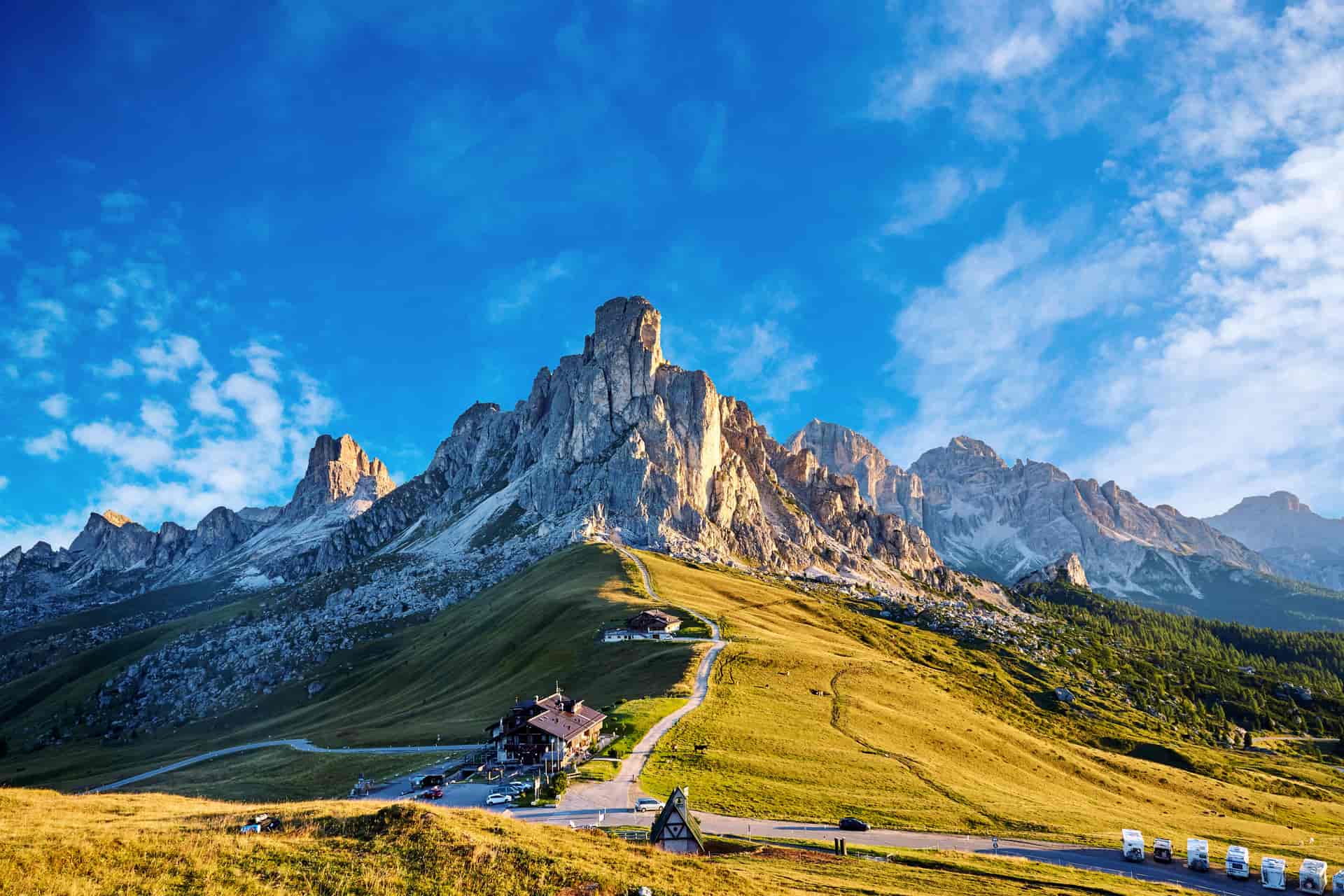 Rugged Dolomite mountains under a bright blue sky with a lodge and winding road in the foreground.