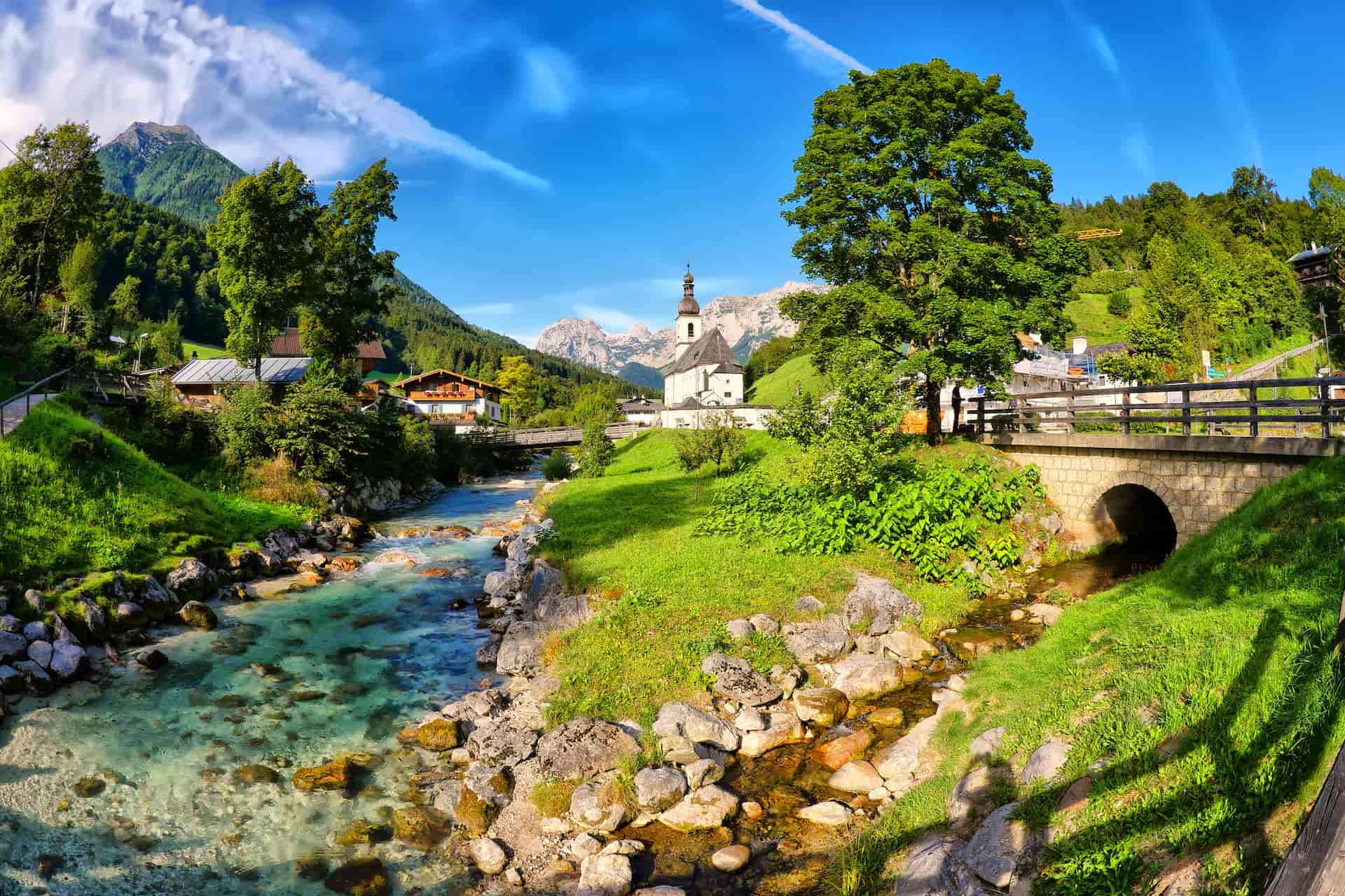Alpine village with clear stream, church, and mountains under a bright blue sky.