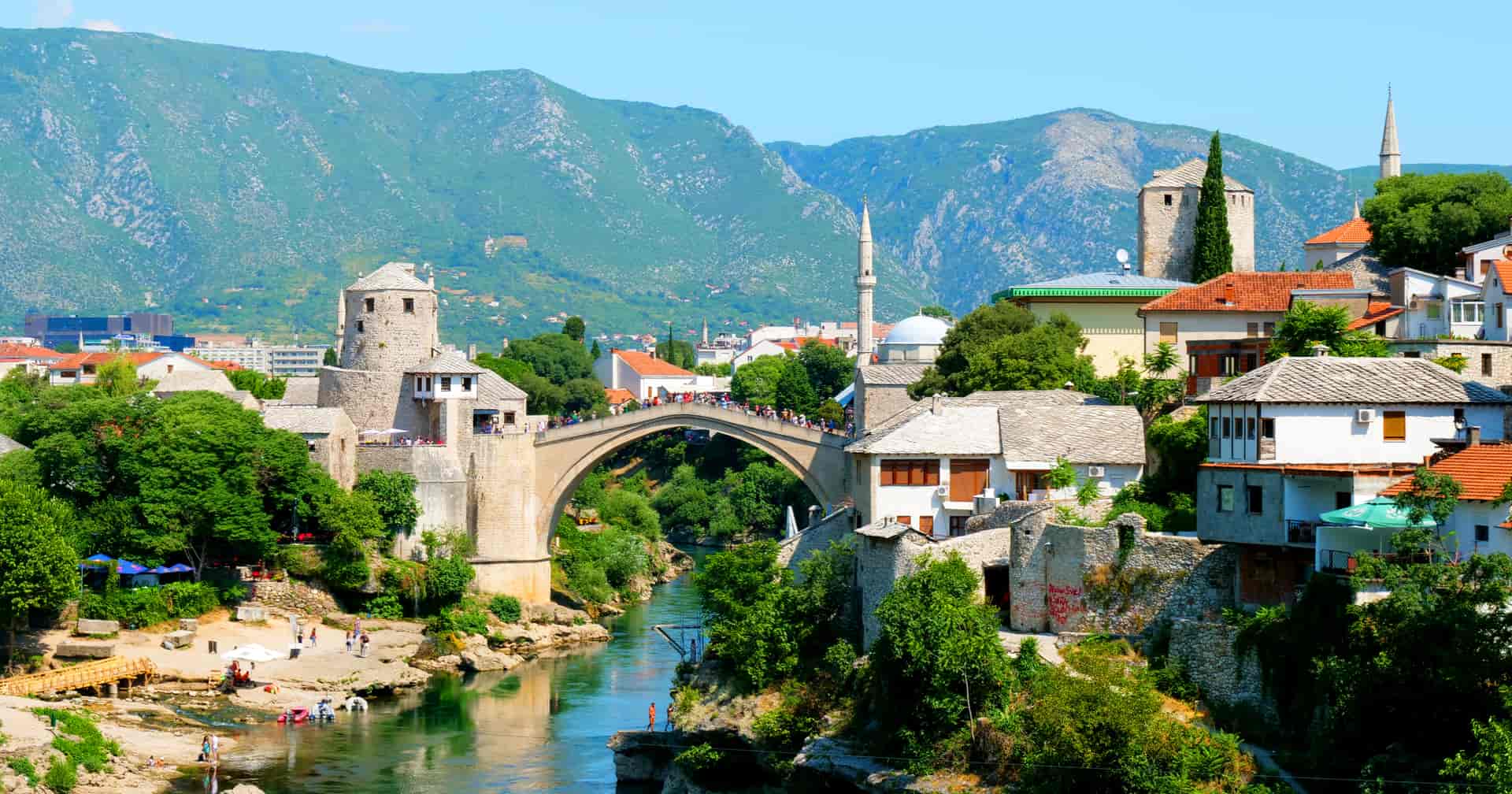 Stone arch bridge over river in Mostar with historic buildings and green mountains.