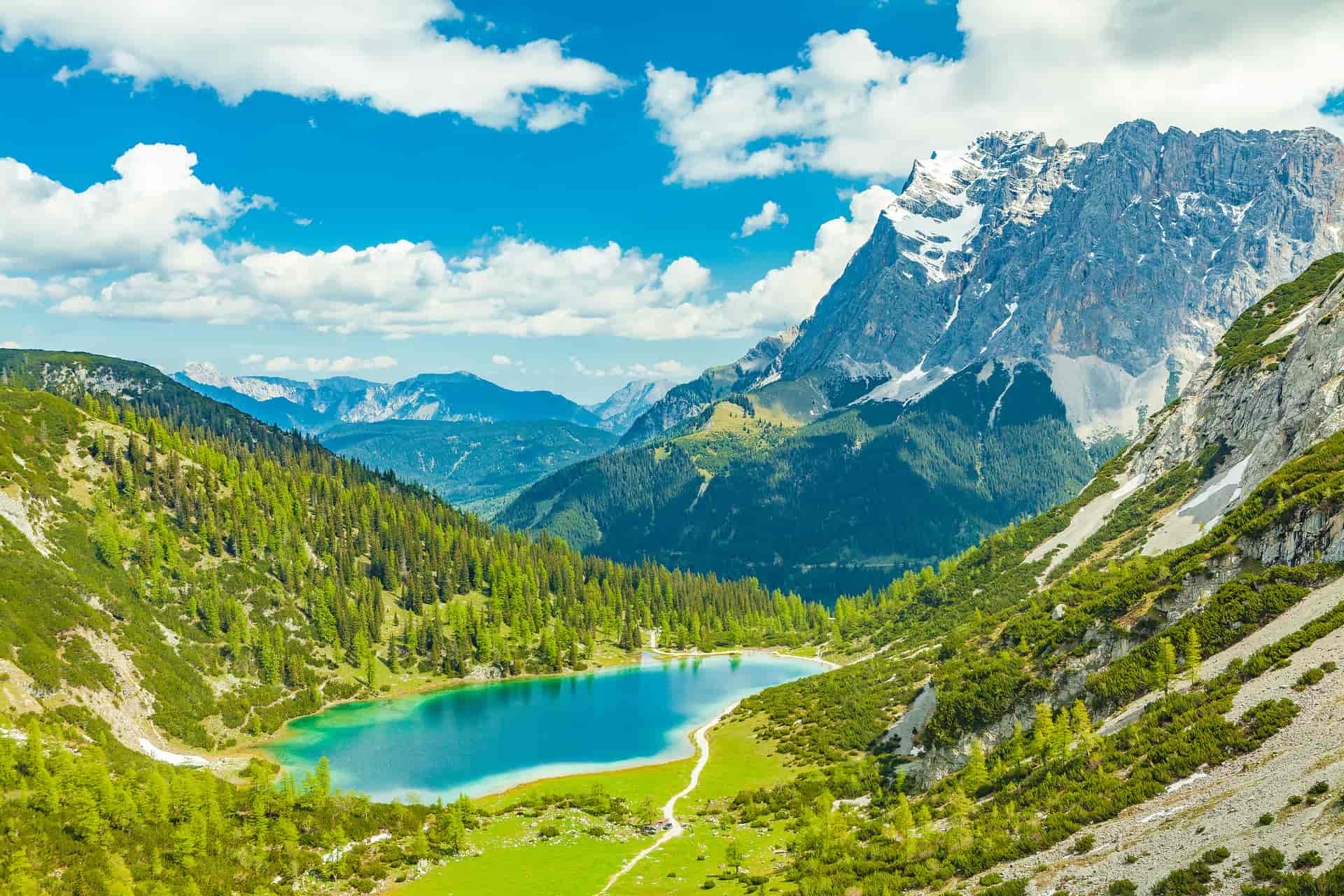 Alpine lake with turquoise water surrounded by green slopes and snow-capped mountains under blue sky.