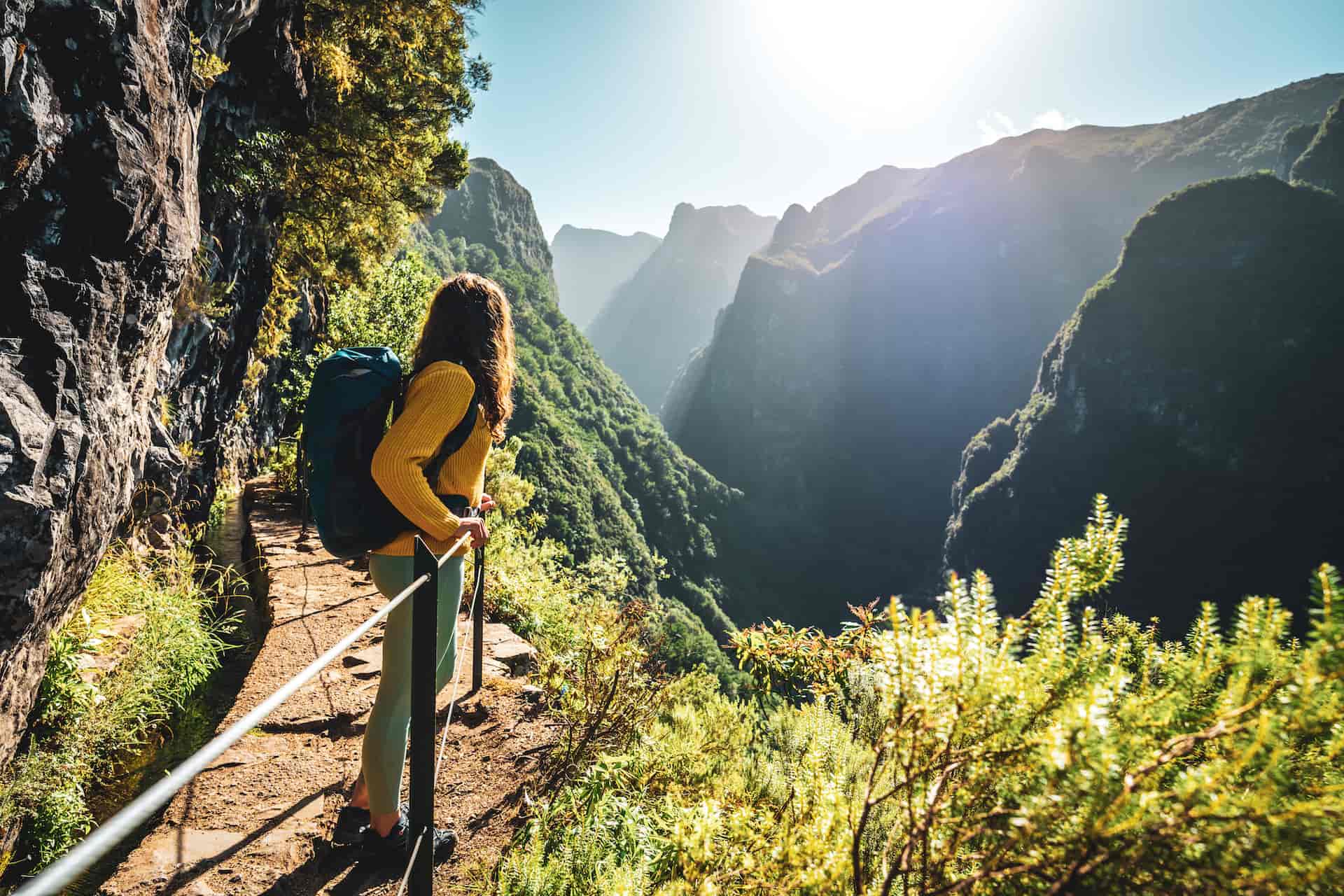 Hiker with backpack on narrow trail overlooking lush, sunlit mountain valley in Madeira.