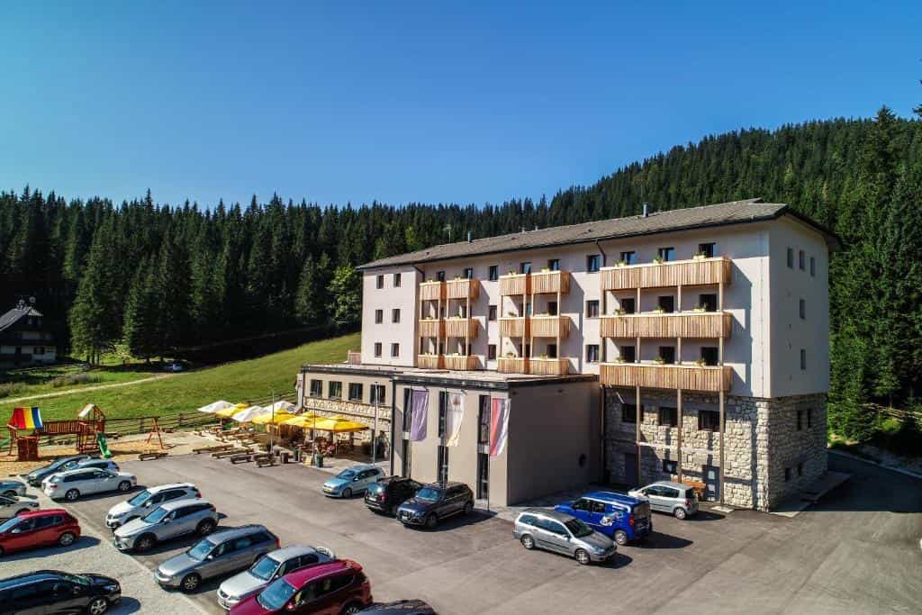 Hotel Pokljuka building with wooden balconies, parking lot, and dense pine forest backdrop.
