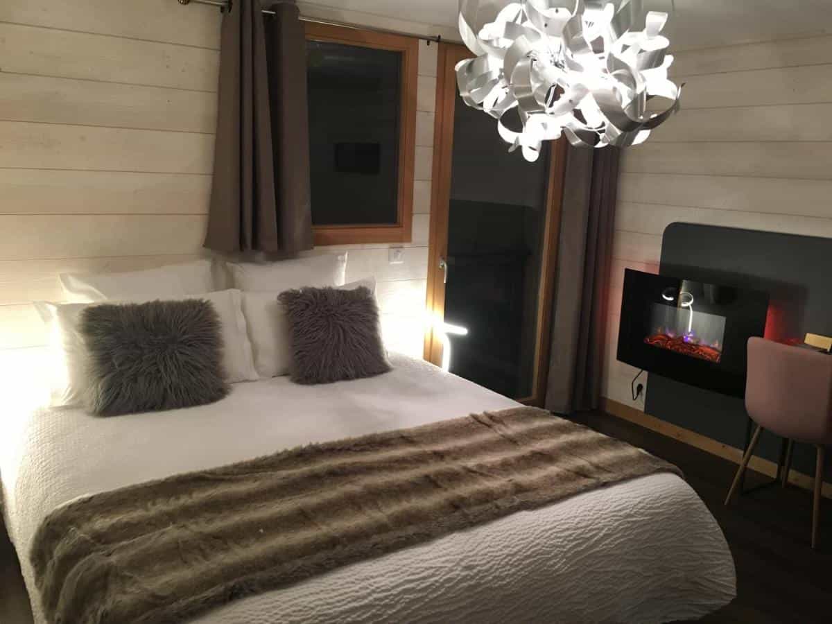 Cozy bedroom with white bedding, faux fur pillows, modern light fixture, and wall-mounted electric fireplace.