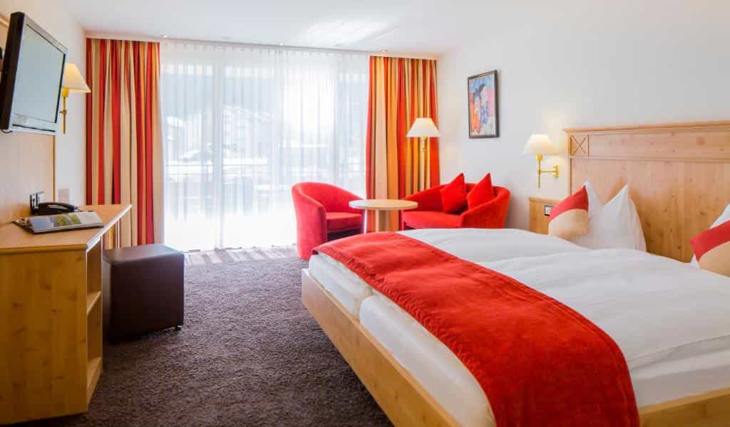 Hotel Taescherhof room with wooden bed, red accents, and balcony view through sheer curtains.