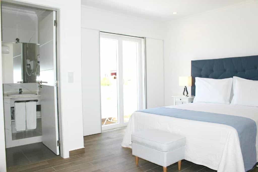 Bright hotel room with white bedding, blue headboard, and open bathroom entrance.