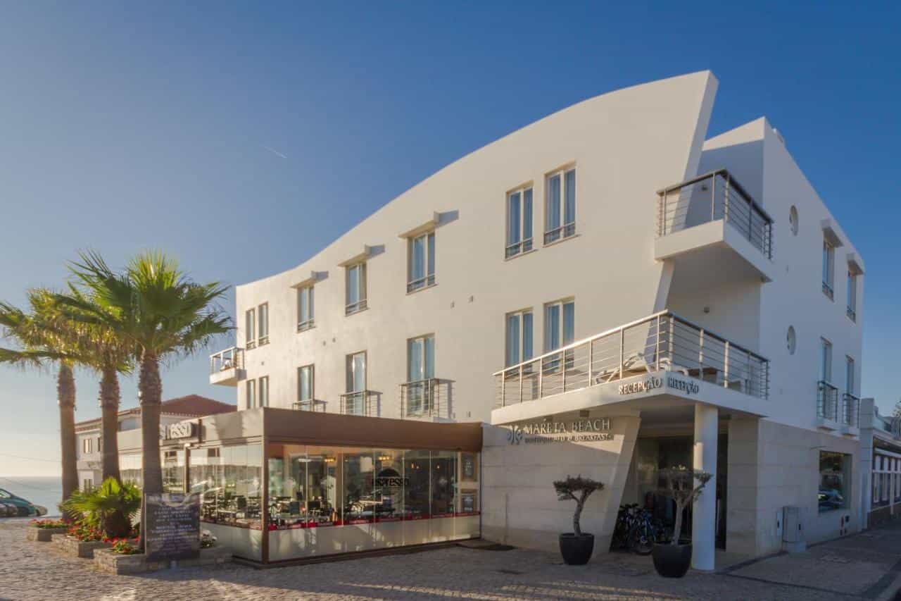 White modern hotel building with "Mareta Beach" sign, palm trees, and cafe under clear blue sky.