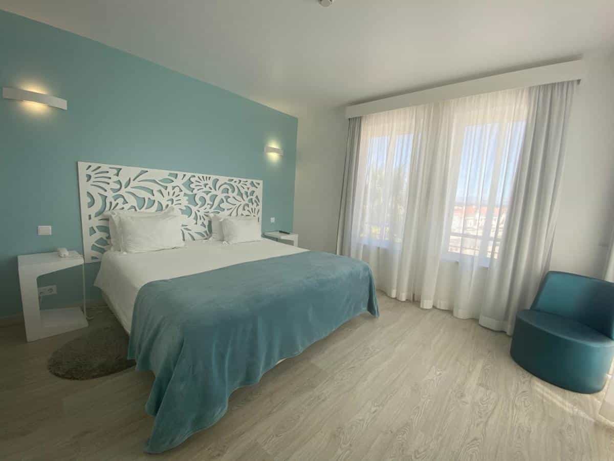 Modern hotel bedroom with teal accent wall, white patterned headboard, and large window with sheer curtains.