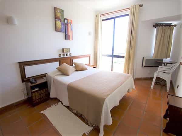 Hotel bedroom with white walls, terracotta tile floor, and a large window with bright light.