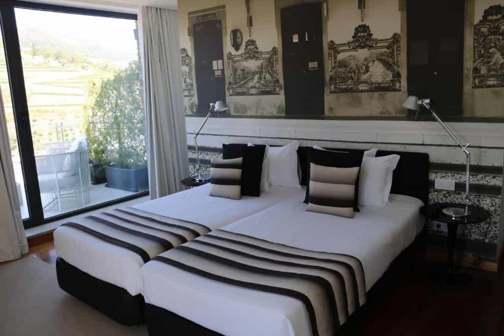 Twin beds in hotel room with balcony view of terraced hillsides and white patio furniture.