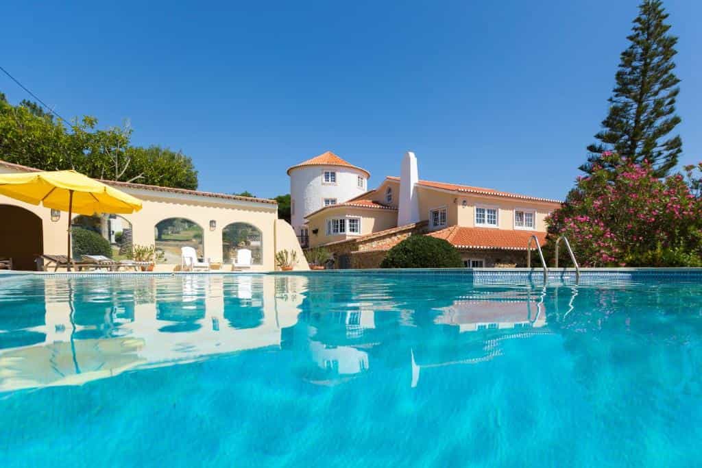 Swimming pool reflecting guesthouse with yellow umbrella and flowering tree under blue sky.