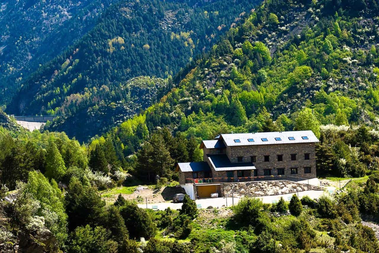 Stone hostel building nestled in lush green mountainside with a distant dam visible