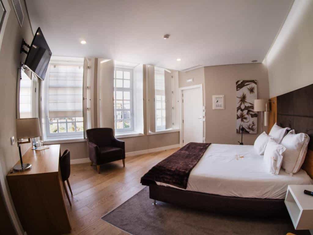 Hotel room interior with large bed, wooden desk, armchair, and windows with white shutters