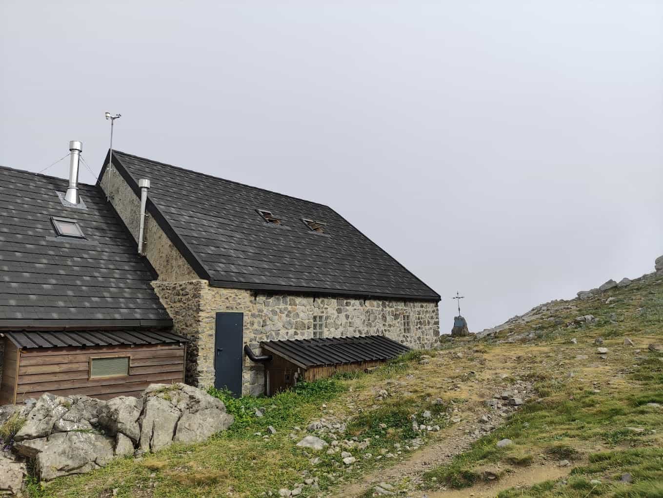 Stone mountain refuge with dark roof next to rocky path under overcast sky, Refugio Collado Jermoso