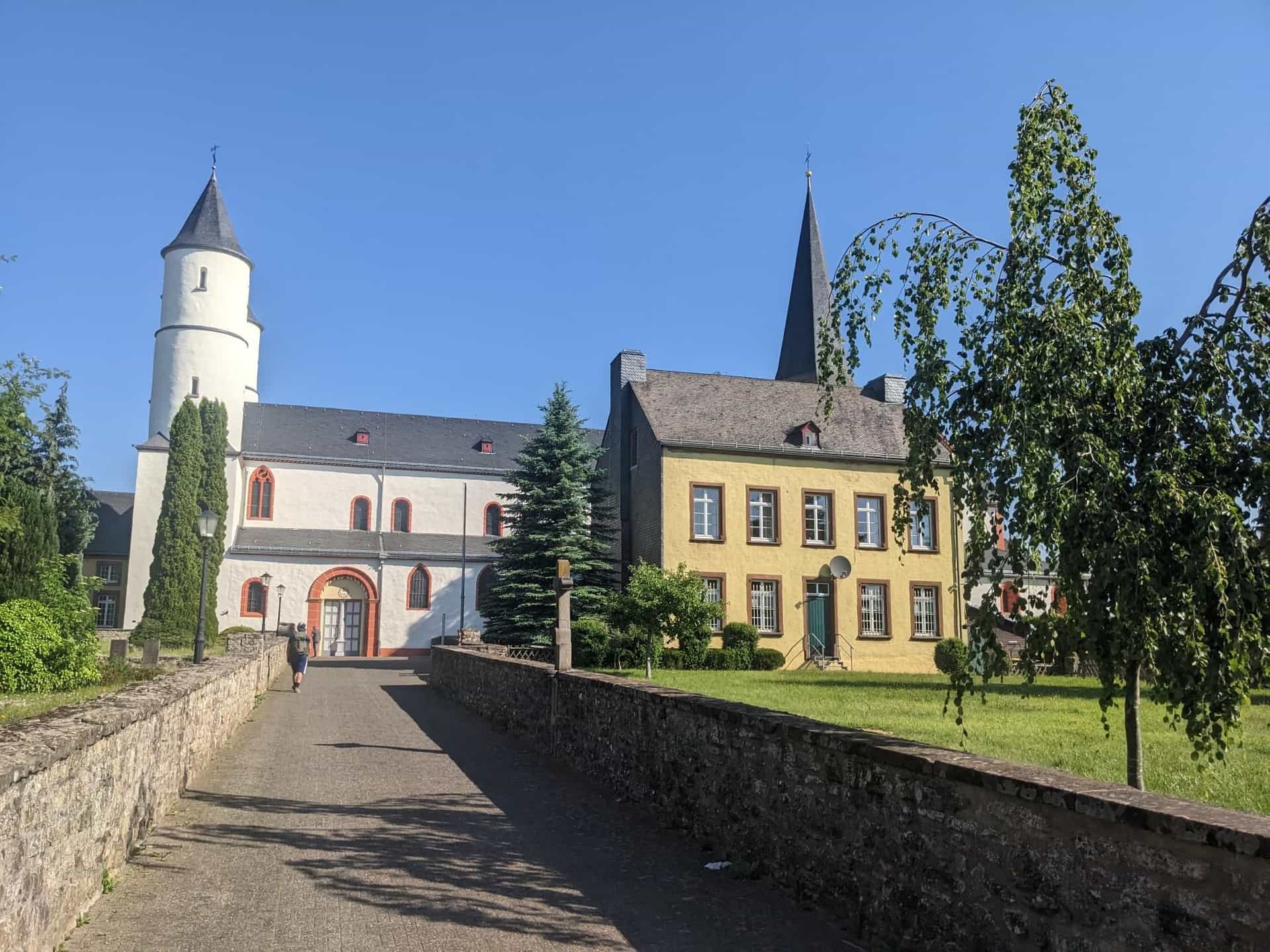 Stone path leading to Kloster Steinfeld church with white and yellow buildings under clear blue sky.