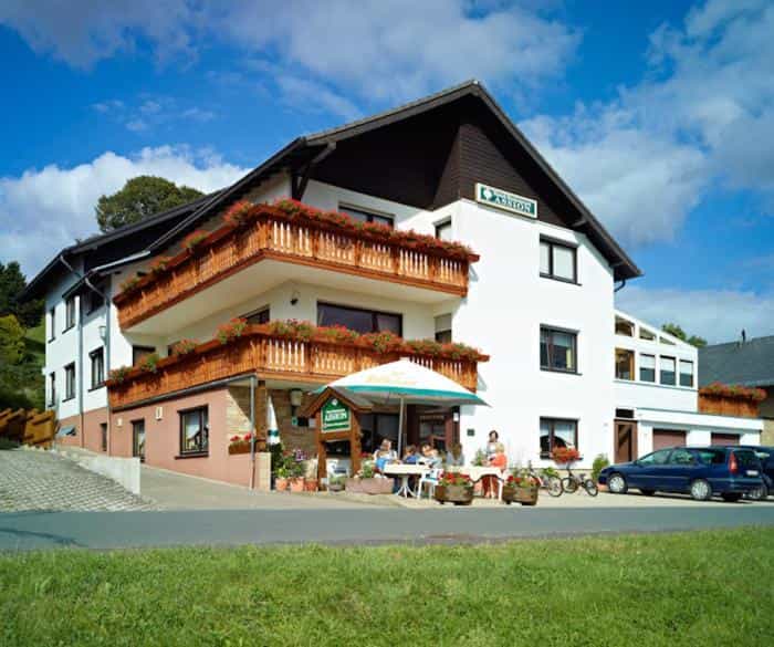 Hotel Garni Assion building with wooden balconies and outdoor dining area under a sunny sky.