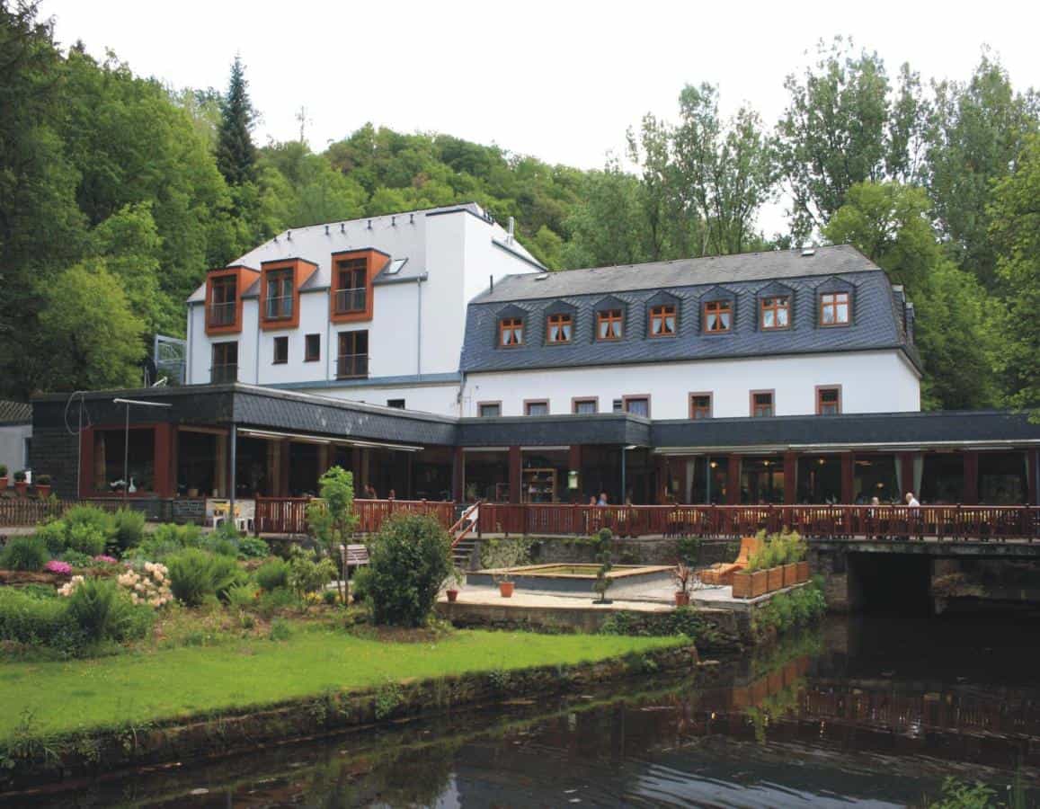 White building with dark roof next to a river, surrounded by lush green forest.