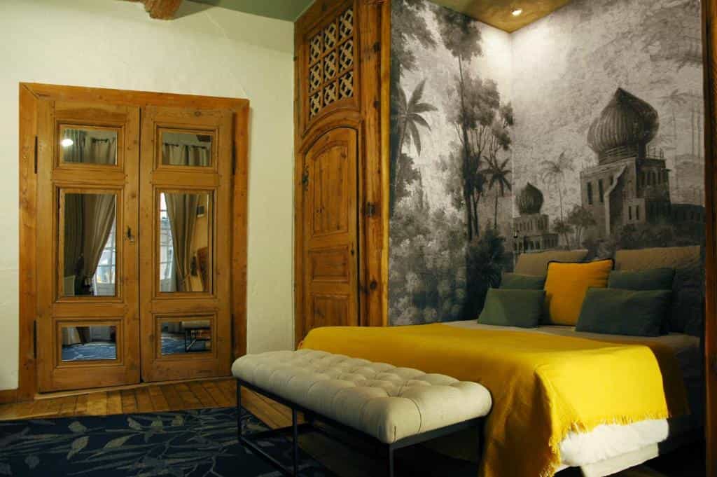 Bedroom with yellow bedding, tufted bench, and exotic mural wallpaper near wooden doors.