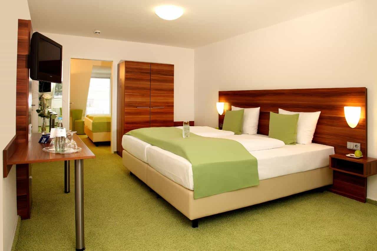 Modern hotel room with large bed, green carpet, wooden furniture, and view into an adjoining room.