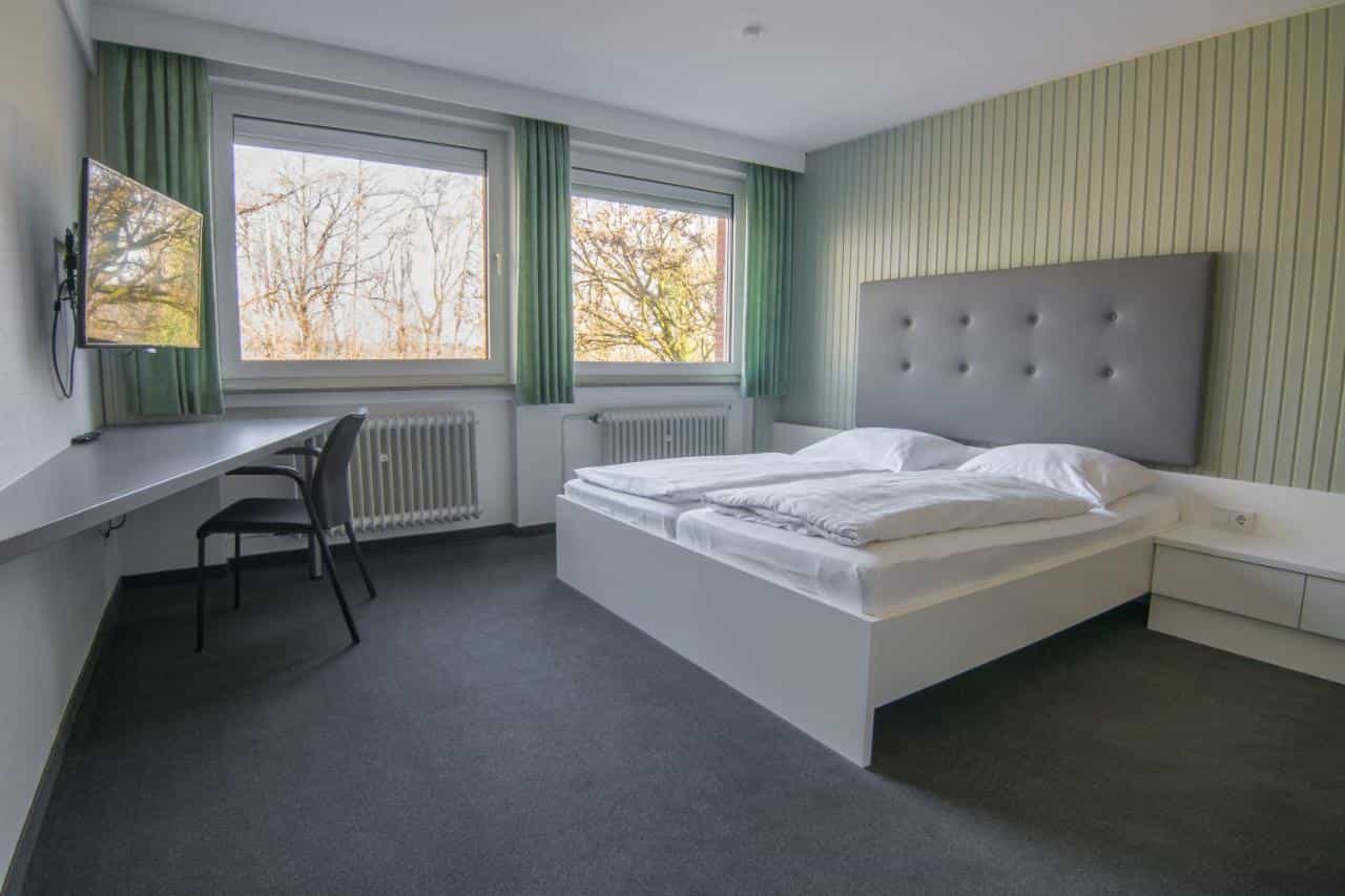 Modern hotel room with white bed, desk, and view of bare trees outside windows.