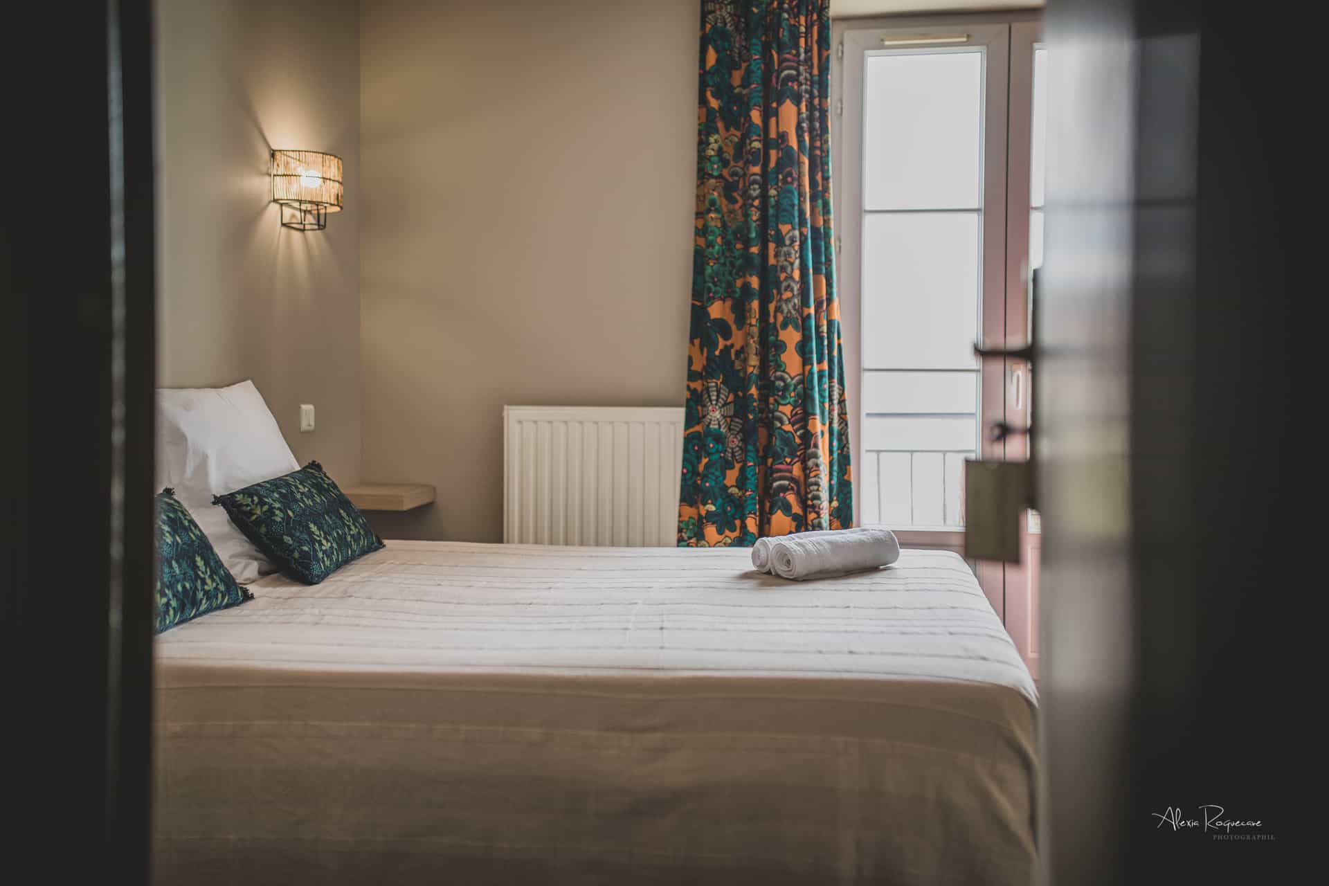 Bedroom with neatly made bed, patterned pillows, and bright floral curtains near a window.