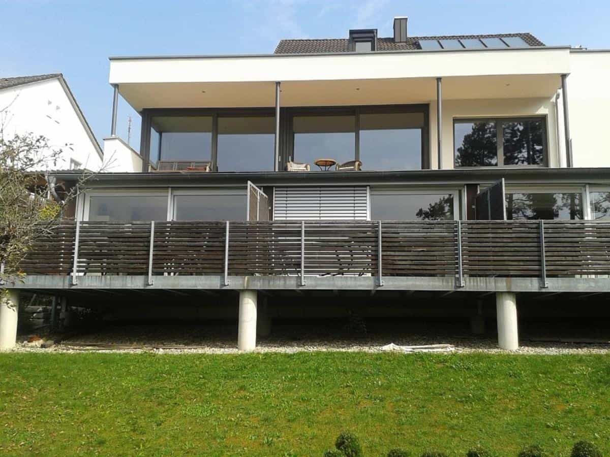 Modern house with large windows and a wooden-railed elevated balcony overlooking a lawn.