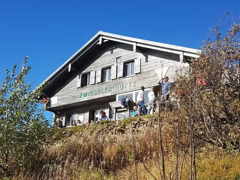 Wooden ZWIESELER HÜTTE mountain hut with people on the terrace under a clear blue sky.