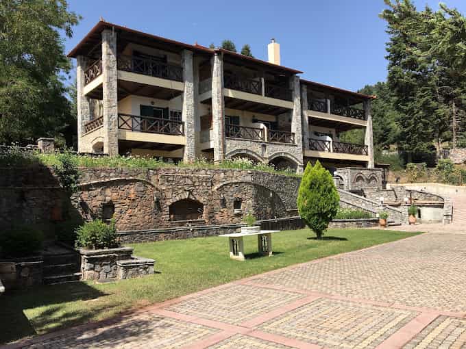 Stone chalet with balconies above a grassy area and paved courtyard, surrounded by trees.