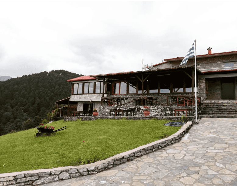 Stone building with Greek flag outside, overlooking dense forest on a cloudy day.