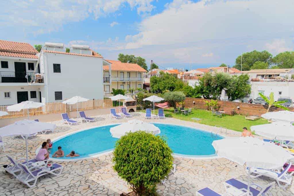 Swimming pool area with sun loungers and white umbrellas beside white apartment buildings under blue sky.