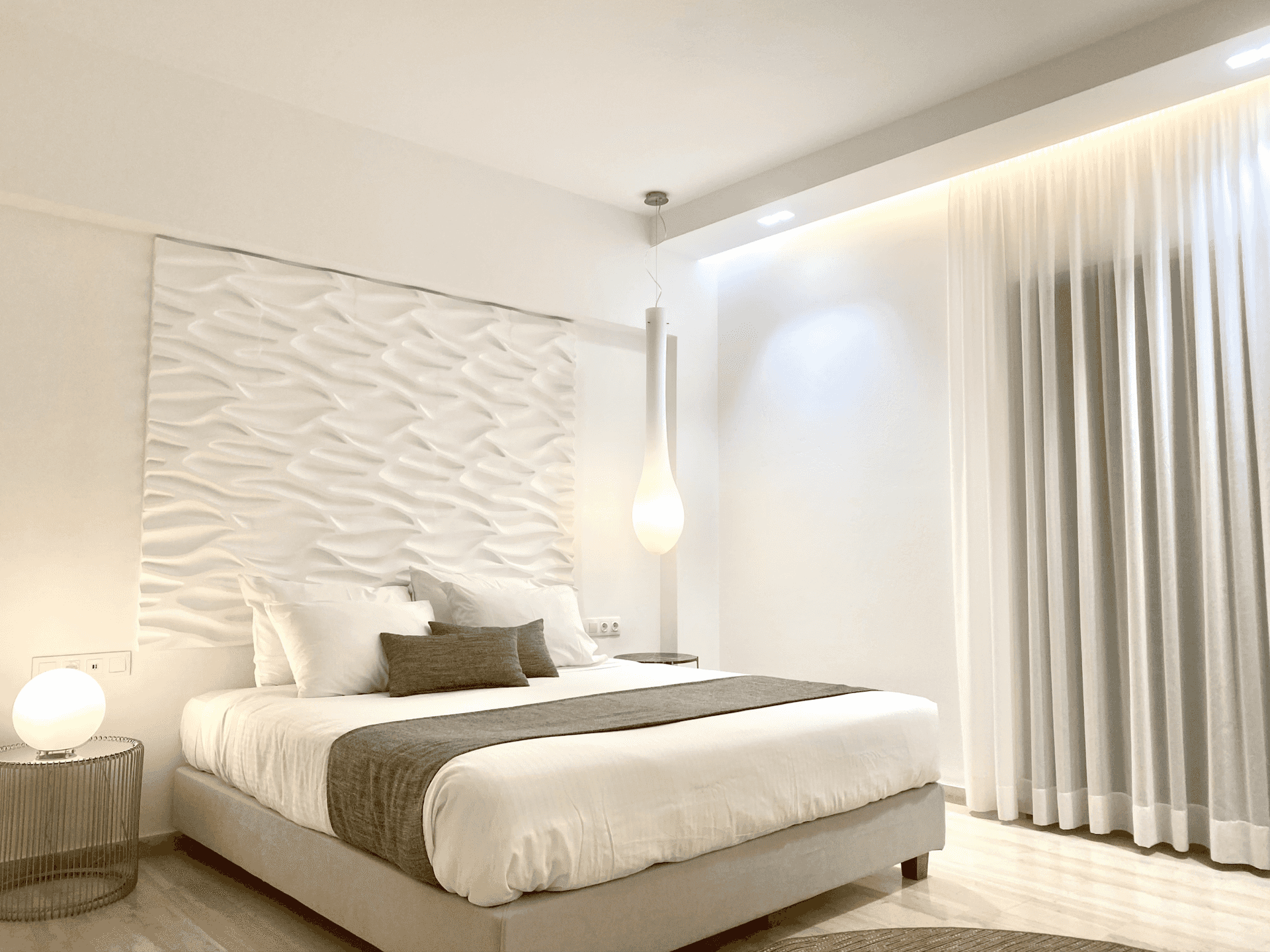 Modern hotel bedroom with white textured headboard, gray bedding, and sheer curtains.
