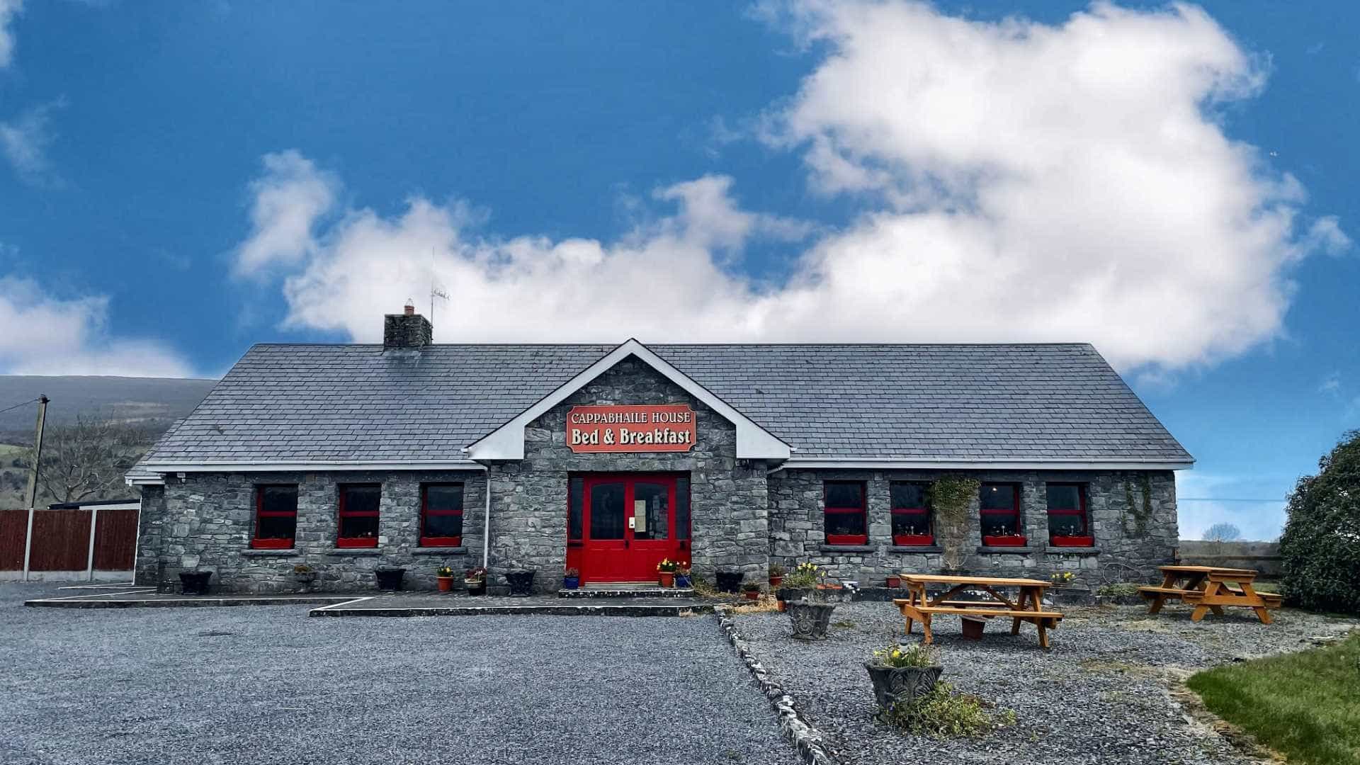 Cappabhaile House Bed & Breakfast with stone facade, red door, and picnic tables under blue sky.