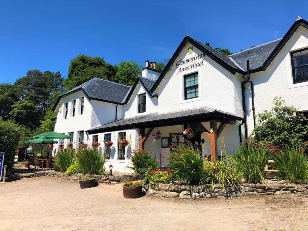 Glenmoriston Arms Hotel, a white building with dark trim, surrounded by lush greenery and flowers.