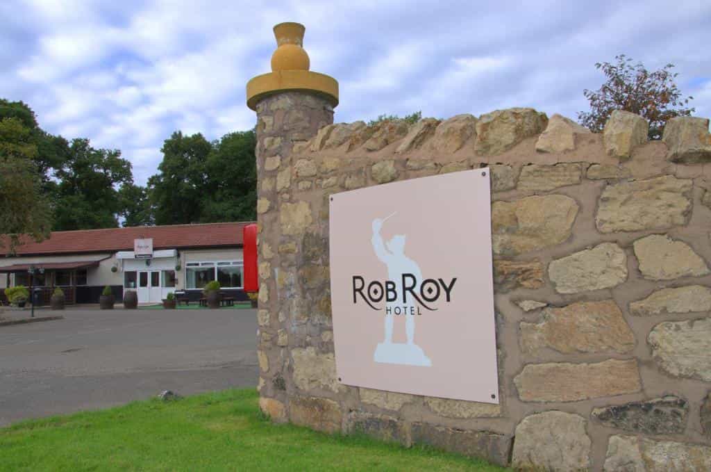 Rob Roy Hotel sign on stone wall with parking lot and low building in background