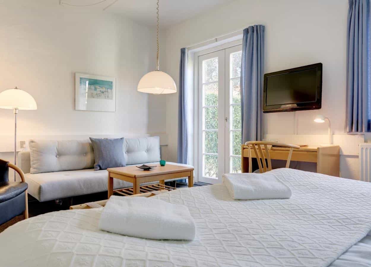 Hotel room with white bedding, light sofa, wooden desk, and balcony doors at Hotel Siemsens Gaard