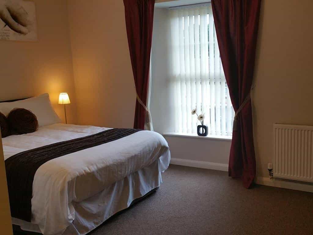 Bedroom with double bed, bedside lamp, window with vertical blinds, and red curtains at The Begelly Arms Hotel.