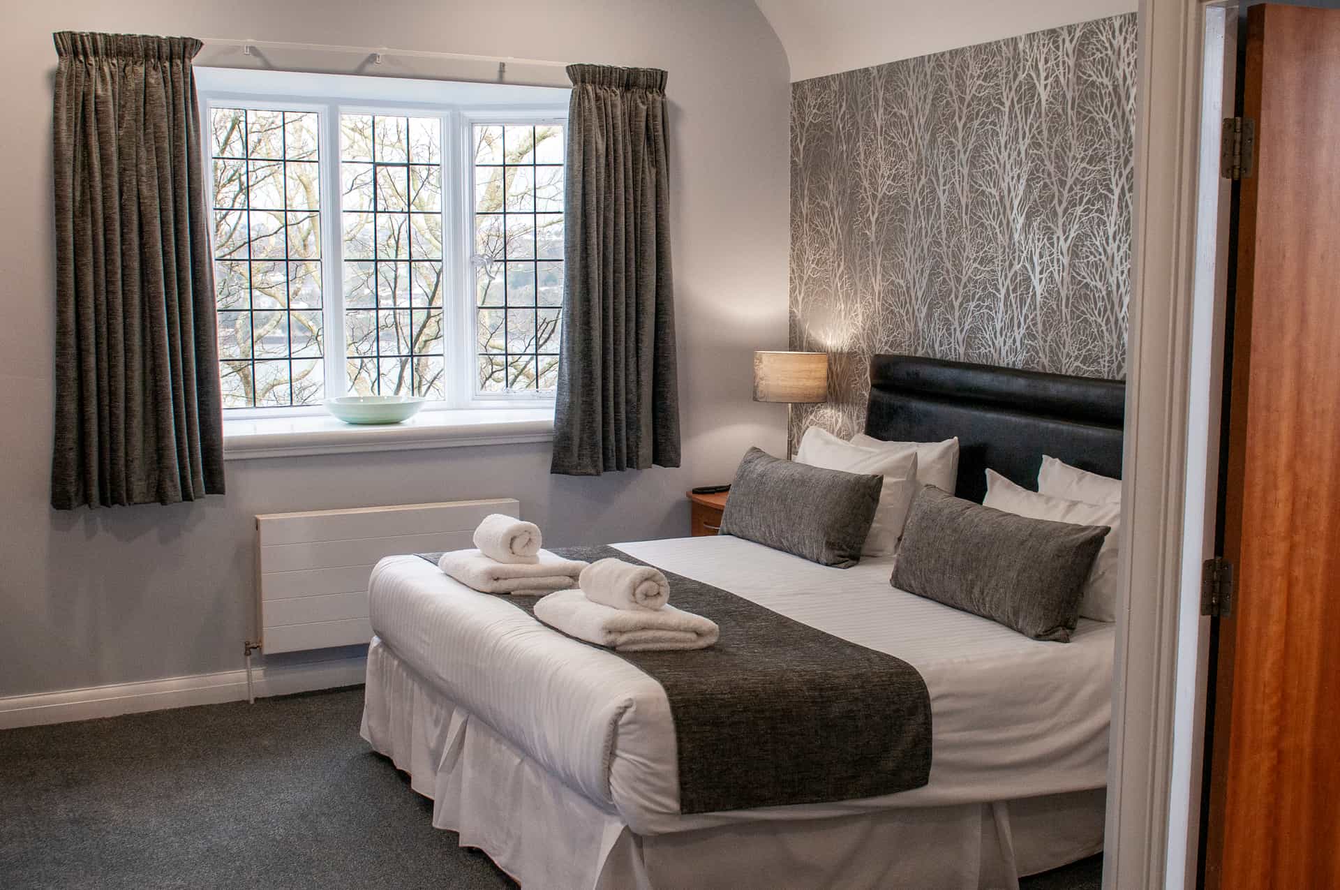Hotel bedroom with patterned wallpaper, bay window view of bare trees, and neatly folded towels.