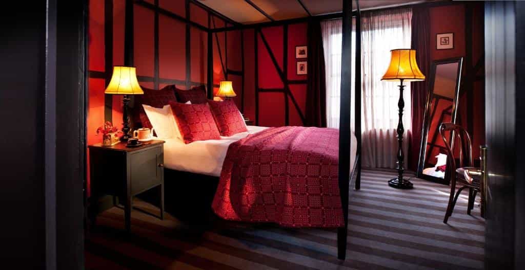 Four-poster bed with red bedding in a room with dark wood beams and warm lighting.