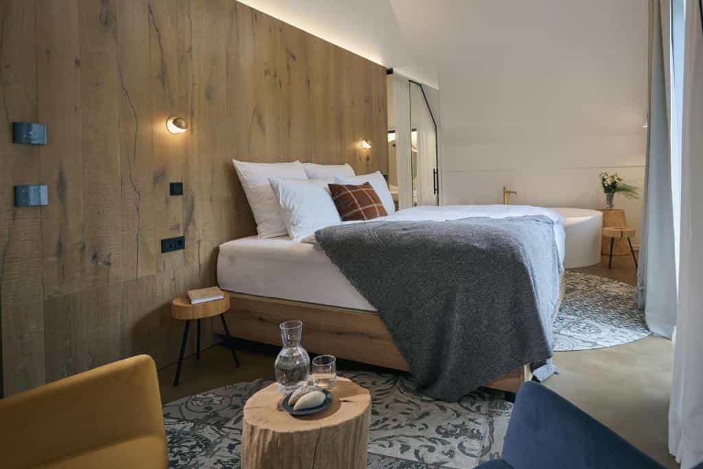 Modern hotel bedroom with wood accent wall, white linens, grey throw, and freestanding bathtub.