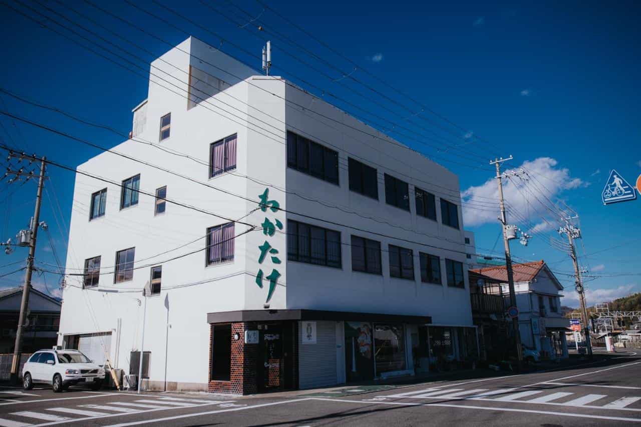 Wakatake Guesthouse, a white building with green Japanese script, on a sunny street corner.