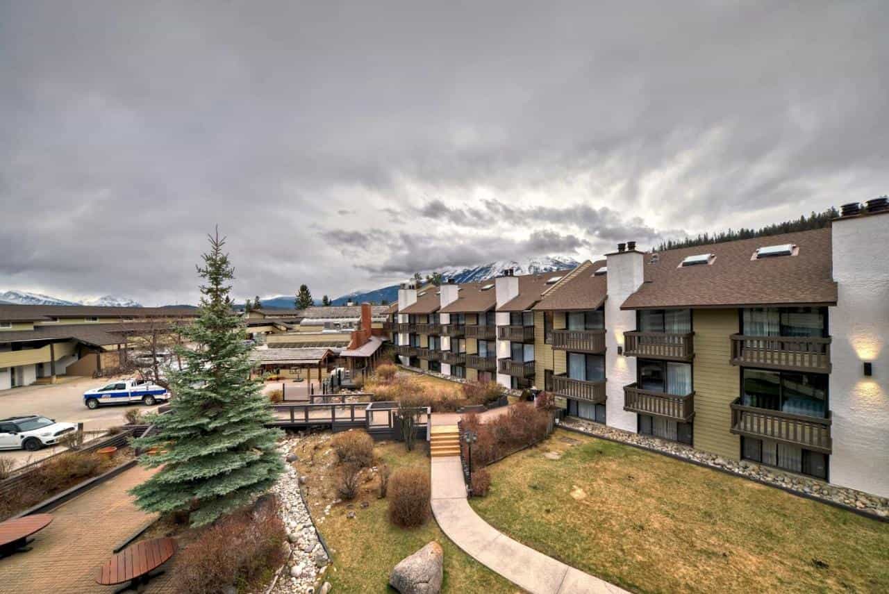 Lodge exterior with balconies, lawn, and snow-capped mountains under cloudy sky.