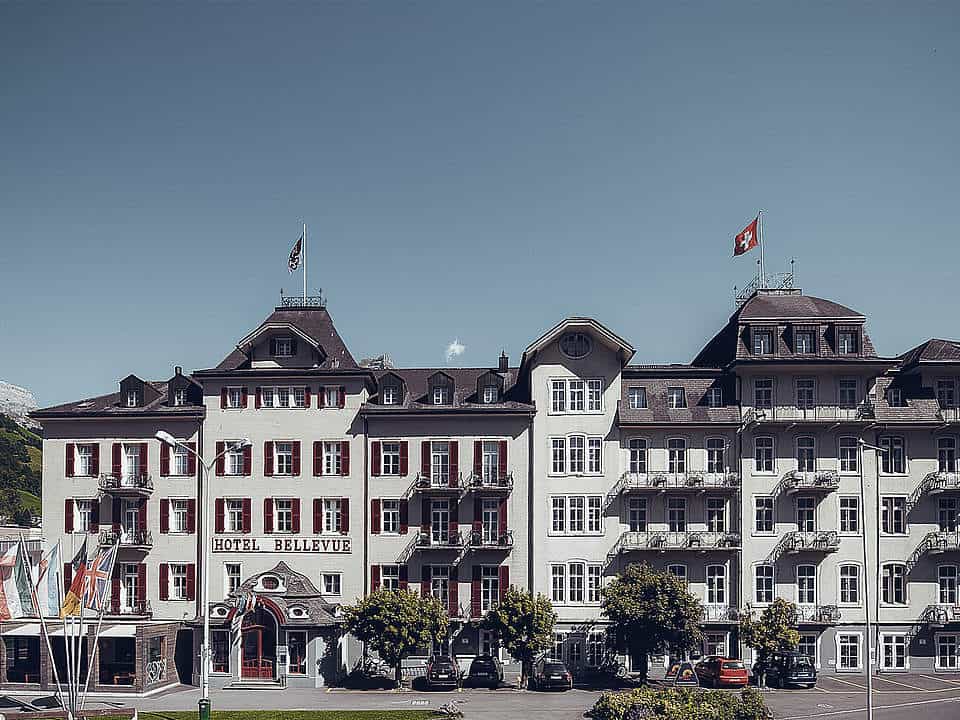 Hotel Bellevue building with Swiss flag flying under a clear blue sky, mountains visible left.