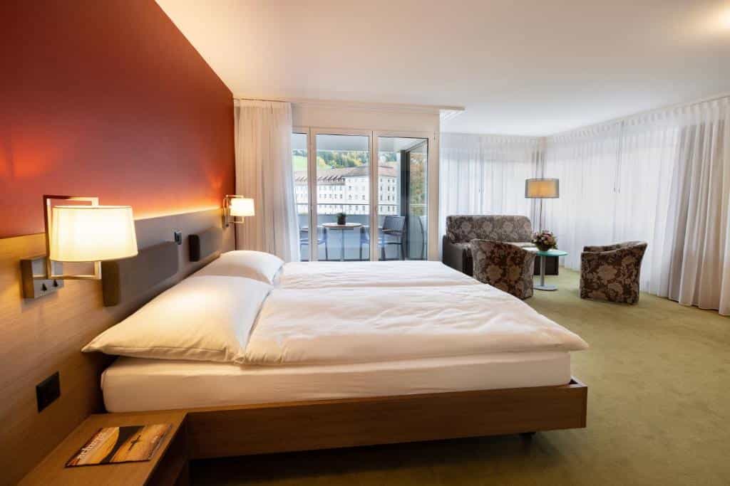 Hotel room with twin beds, seating area, and balcony view of buildings and hills.