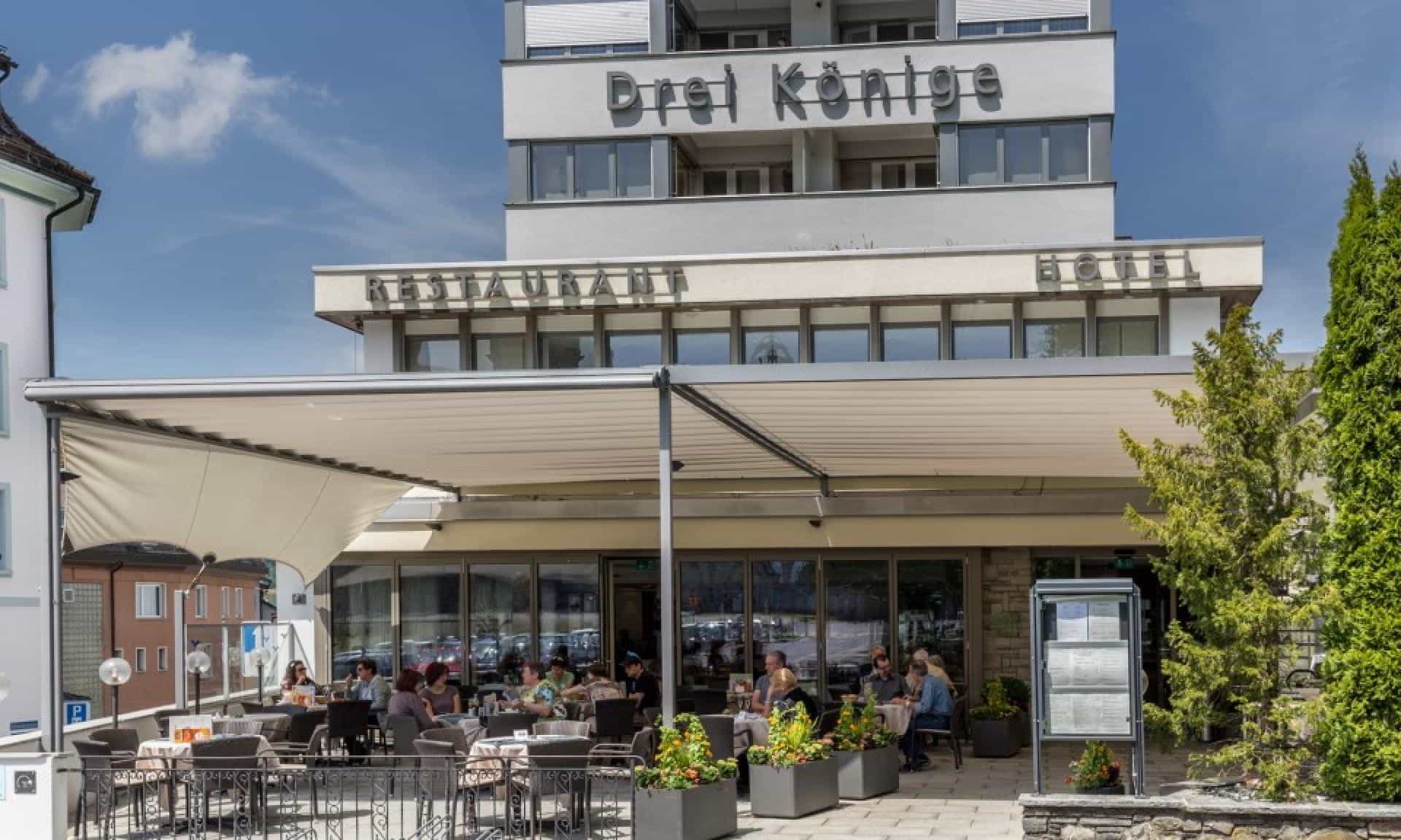 Outdoor restaurant patio dining beneath awning at Hotel Drei Könige building