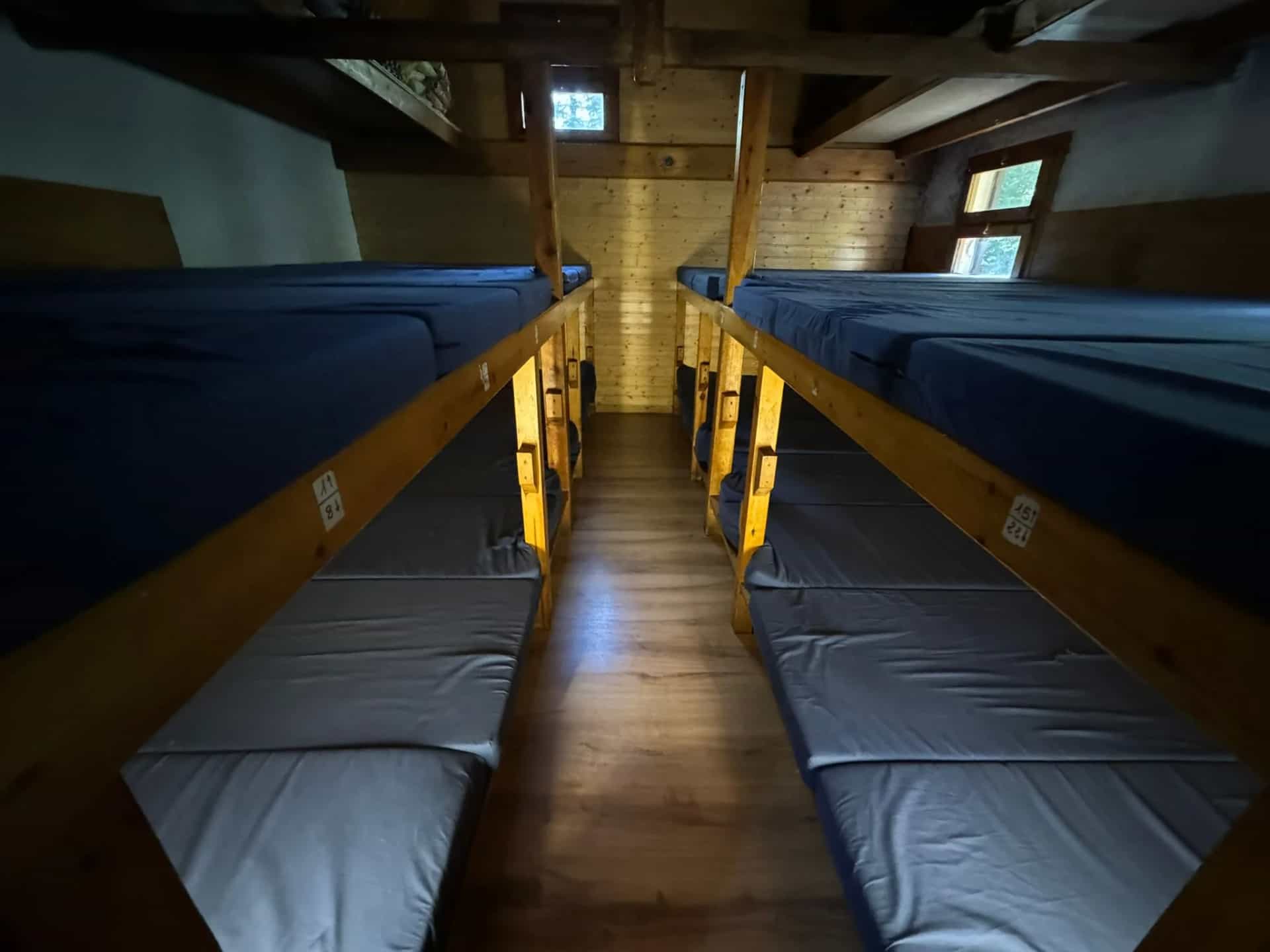Bunk beds with blue mattresses in a rustic wooden dormitory room with small windows.