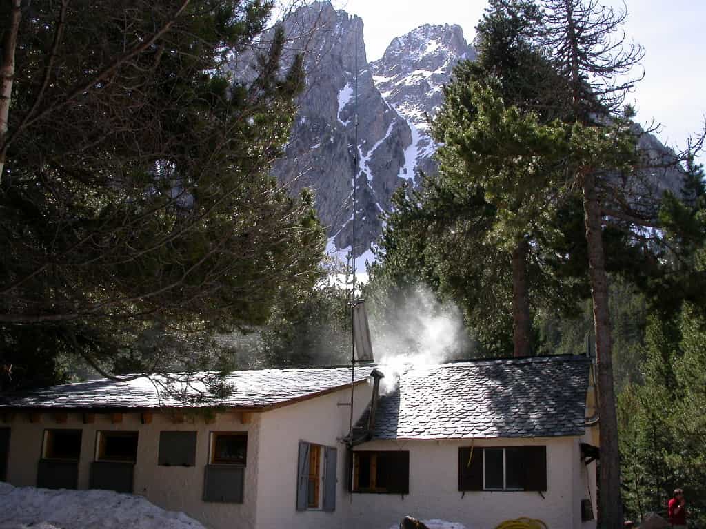 Mountain refuge with smoke rising from chimney, set against snowy peaks and pine trees.