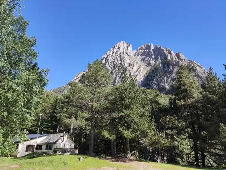 Mountain refuge hut nestled among pine trees below a rugged, rocky peak under a clear blue sky.