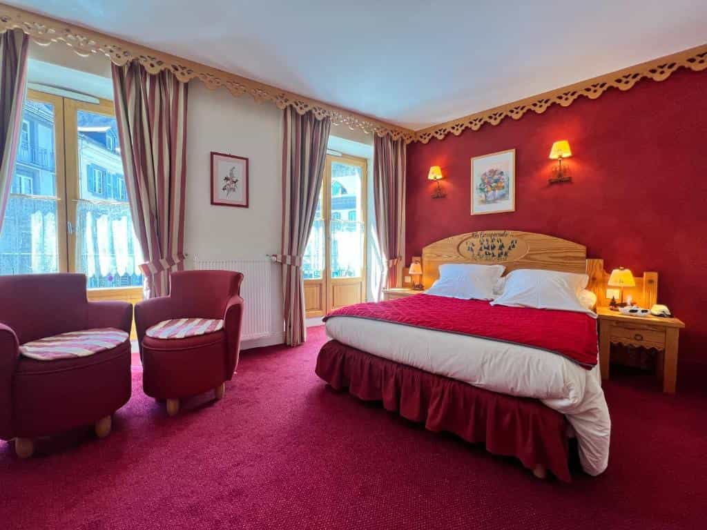 Hotel bedroom with red carpet, wooden headboard, and two red armchairs near windows.