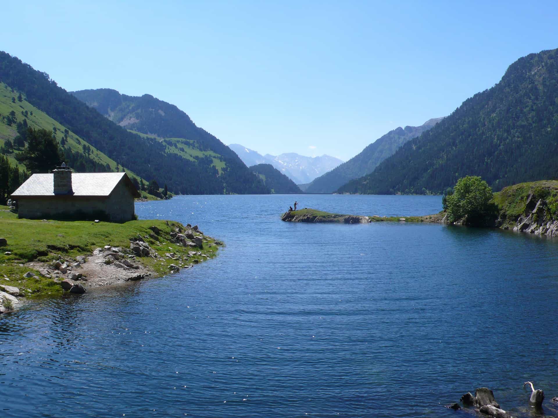Stone cabin by blue alpine lake with mountains and people fishing on a small island.