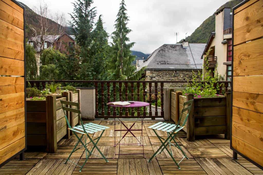 Balcony with wooden deck, metal table, and chairs overlooking mountain village buildings.