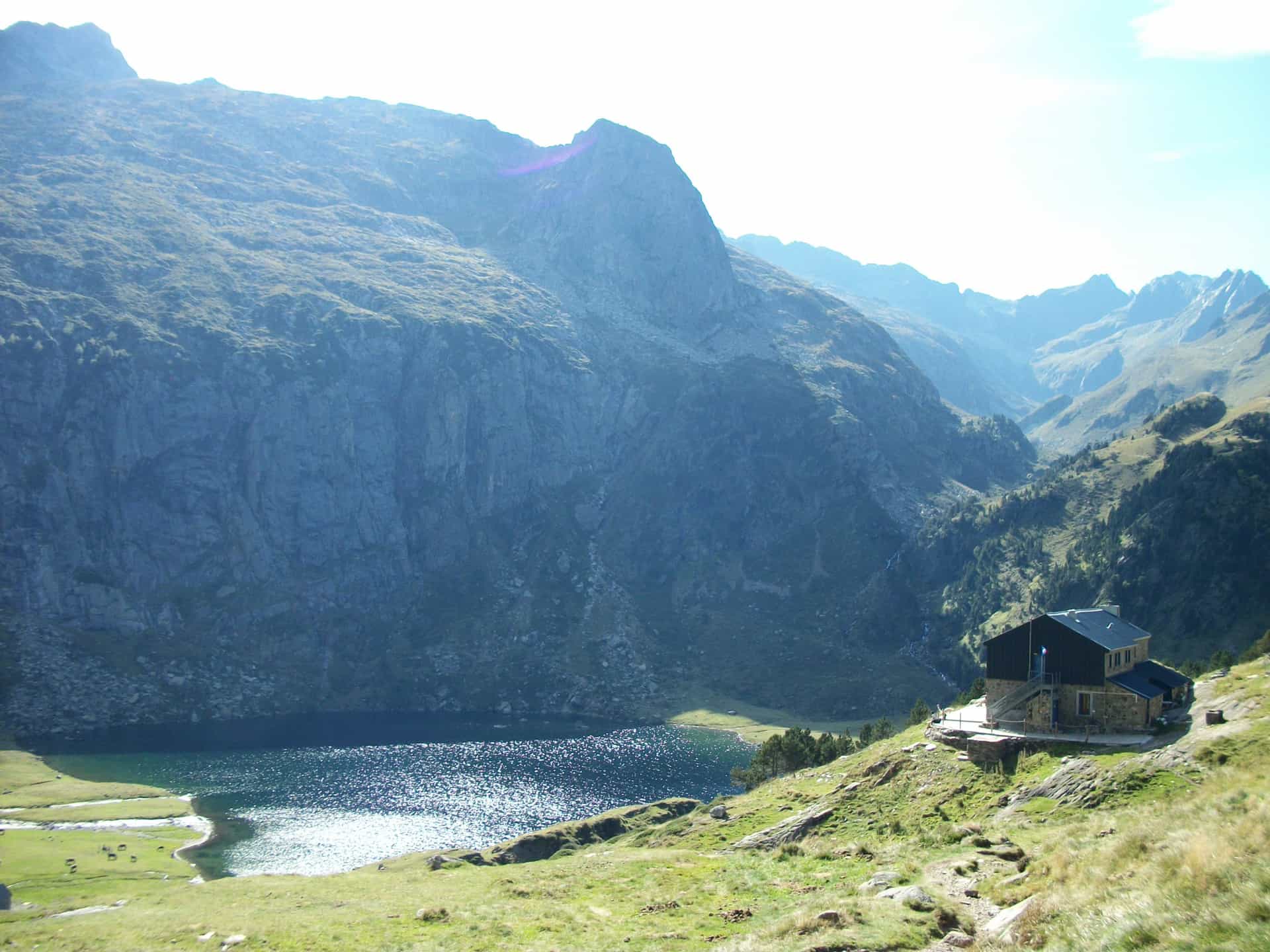 Stone refuge building near a sparkling alpine lake beneath large, sunlit mountains.